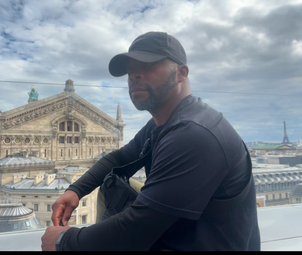 E.N. Howie seated on a Paris rooftop with the Palais Garnier in the background and the Eiffel Tower in the distance, wearing black clothing and a cap, reflecting in thought.